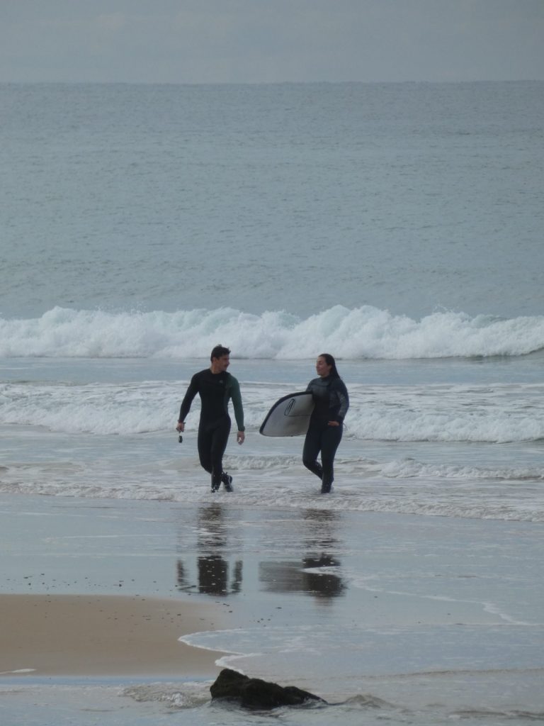 Dos surfistas con traje de neopreno caminando por la orilla en un día nublado tras surfear , Clima y rendimiento en surf es clave